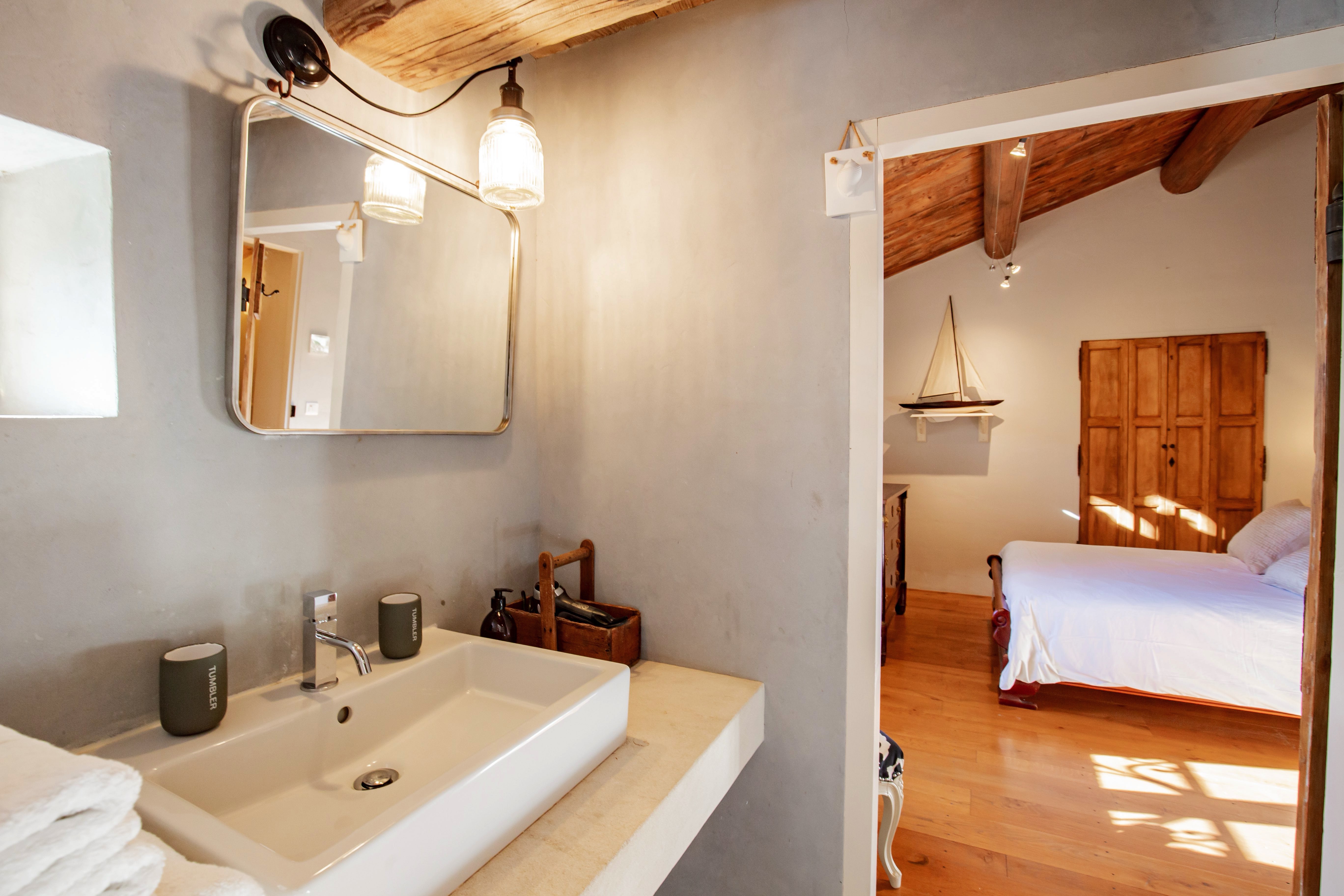 A cozy bathroom with a large rectangular sink, wooden accents, and a small bedroom visible through an open door, featuring warm lighting and a comfortable, inviting atmosphere.